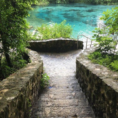 Stone steps lead down to a clear blue water body, surrounded by lush greenery and trees.