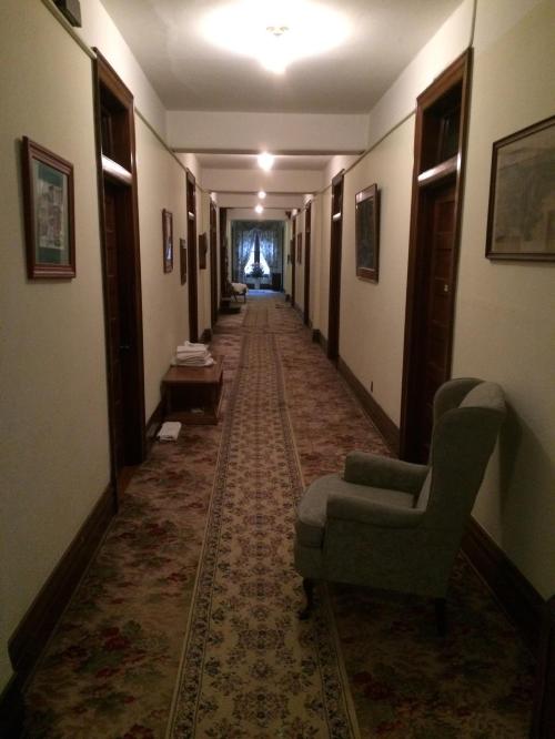 A long, dimly lit hallway with wooden doors, framed pictures, and a patterned carpet, leading to a window at the end.