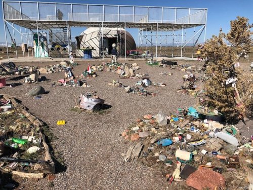 A landscape littered with trash, featuring scattered debris and a modern structure in the background under a clear sky.