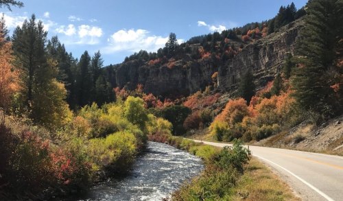 A scenic view of a winding road alongside a river, surrounded by vibrant autumn foliage and rocky cliffs.