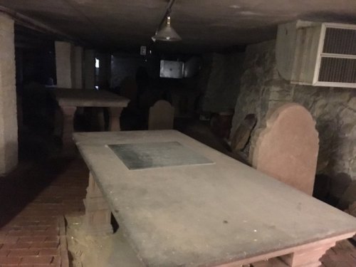 Dimly lit basement with stone tables and chairs, featuring exposed brick and a stone wall.