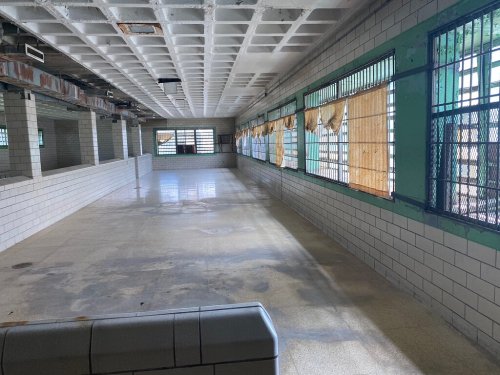 Abandoned interior of a building with tiled walls, empty space, and barred windows, featuring sunlight streaming in.