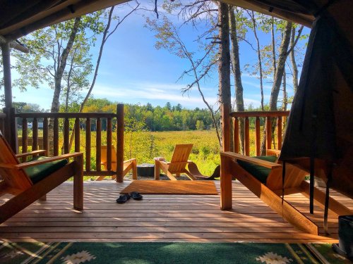 View from a tent with wooden chairs overlooking a grassy field and trees under a clear blue sky.