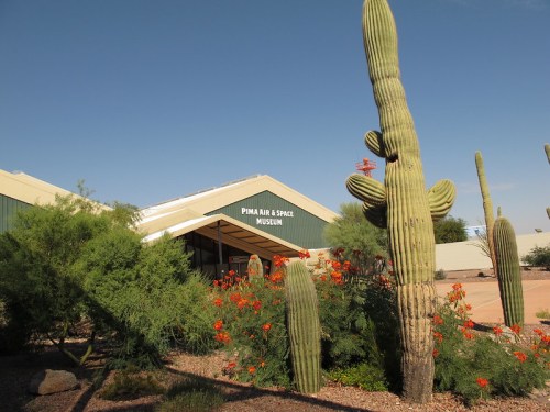 Exterior of the Pima Air & Space Museum, featuring cacti and colorful flowers under a clear blue sky.