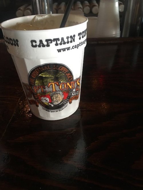 A cup from Captain Tony's, featuring a logo and a drink with a straw, placed on a dark wooden bar surface.