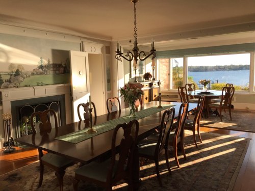A sunlit dining room with a large table, elegant chairs, and a view of a lake through large windows.