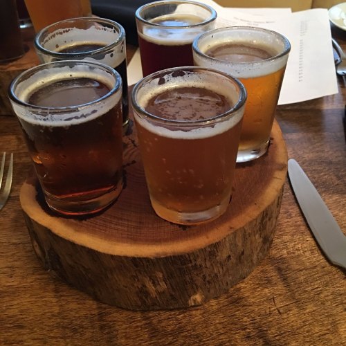 A wooden platter holds five small glasses of different beers, with a rustic table in the background.