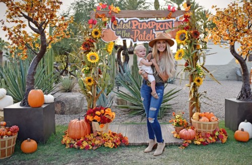 A woman holds a baby in front of a pumpkin patch sign, surrounded by pumpkins and autumn decorations.