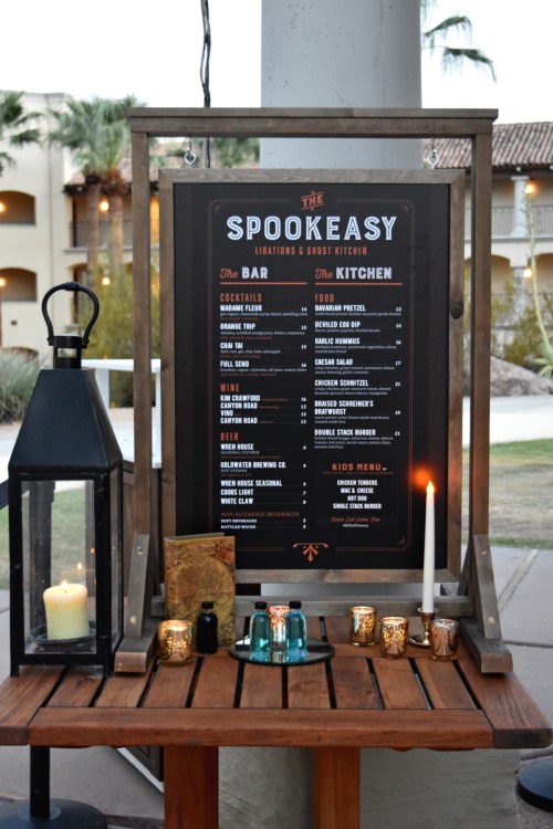 A wooden sign displaying the menu for "The Spookeasy," featuring drinks, food options, and a kids' menu, with candles and lantern.