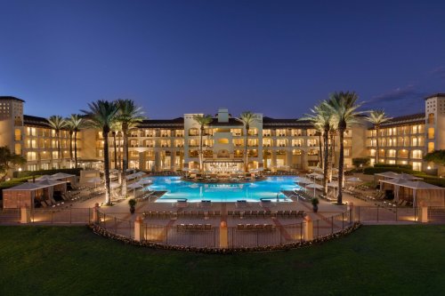 A luxurious hotel at dusk, featuring a large pool surrounded by palm trees and lounge chairs.