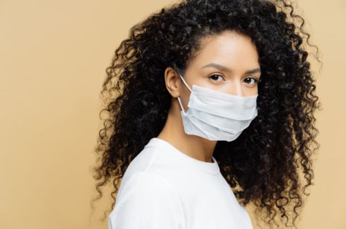 A woman with curly hair wearing a surgical mask, looking over her shoulder against a beige background.