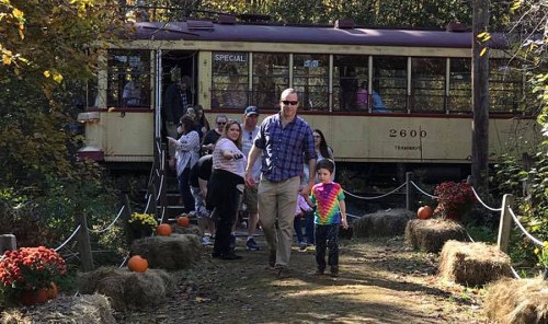A family exits a vintage tram in a fall setting, surrounded by pumpkins and colorful foliage.