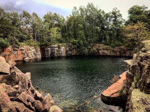 A serene, green-tinted quarry pond surrounded by rocky cliffs and lush trees under a cloudy sky.