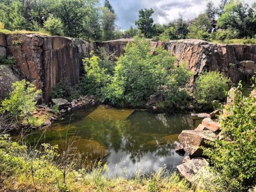 A serene quarry with steep rock walls, lush greenery, and a calm pool reflecting the sky and surrounding trees.