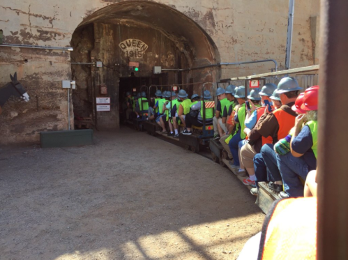 A group of people in hard hats and vests sitting on a mine train, preparing to enter a tunnel marked "Queen 1915."
