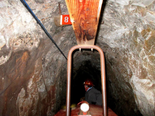 A dimly lit mine tunnel with a wooden beam overhead and a miner in a hard hat near a numbered sign.