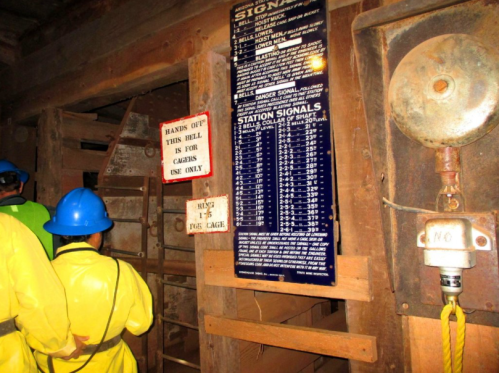 Two people in yellow raincoats and blue helmets stand near a wooden structure with a safety sign and signal instructions.