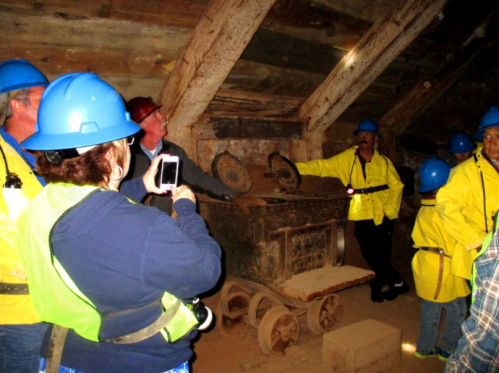 A group of people in hard hats and safety vests examines an old mining cart in a dimly lit underground setting.