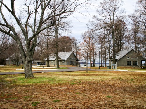 A serene landscape featuring two wooden cabins surrounded by bare trees and a grassy area under a cloudy sky.