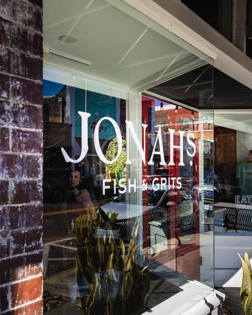 A storefront window displaying the sign "Jonah's Fish & Grits" with reflections of the street and a person nearby.