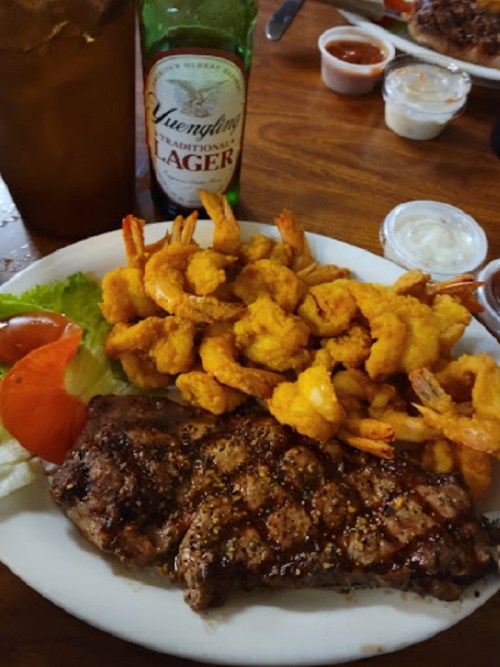 A plate of grilled steak with seasoned shrimp, lettuce, and tomato, alongside a bottle of lager.