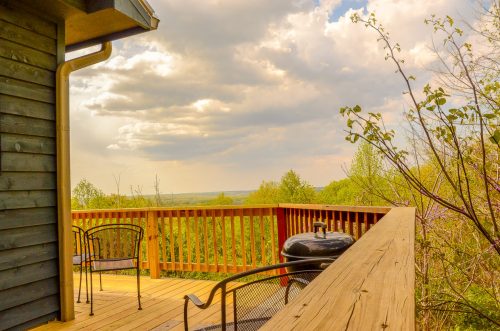 A wooden deck with chairs and a grill overlooks a lush green landscape under a partly cloudy sky.