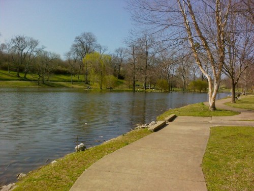 A serene park scene featuring a calm lake, trees, and a paved walkway on a sunny day.