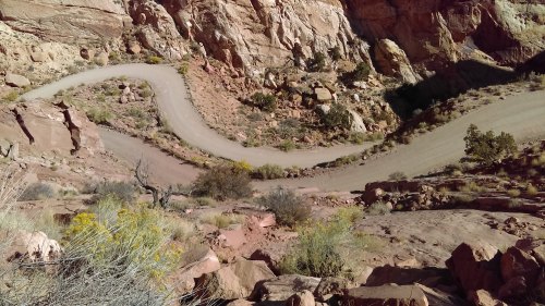 Winding dirt road through rocky terrain with sparse vegetation and steep cliffs in a desert landscape.
