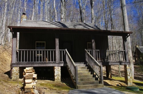 A rustic wooden cabin with a front porch, surrounded by tall trees and stacked firewood in the foreground.