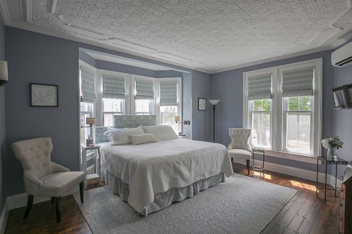 Cozy bedroom with a large bed, two chairs, and bay windows, featuring soft blue walls and elegant decor.