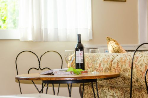 A cozy living space featuring a wooden table with a wine bottle, glasses, and a magazine beside a floral sofa.