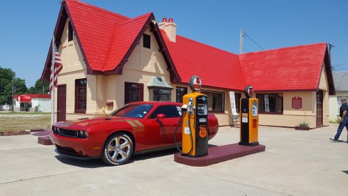 A red sports car parked at vintage gas pumps in front of a charming, red-roofed building on a sunny day.