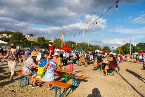 A lively outdoor market scene with people at colorful tables, enjoying food and socializing under a blue sky.