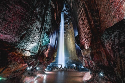 A stunning cave interior with colorful rock formations and a waterfall illuminated by light beams.