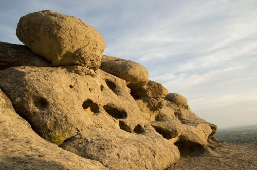Rocky outcrop with large boulders and textured surfaces, set against a clear sky and distant landscape.