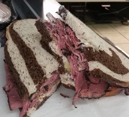 A large, stacked sandwich with layers of pastrami on dark rye bread, partially cut to reveal the filling.