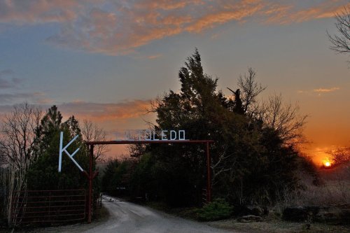 A rustic entrance gate with the letter "K" at sunset, surrounded by trees and a gravel road leading into the distance.