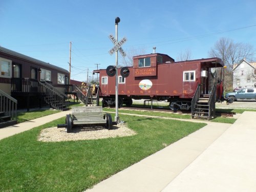 A red caboose on display near a bench and railroad crossing sign, with green grass and blue sky in the background.