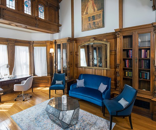 A cozy, well-lit room featuring wooden walls, a blue sofa, chairs, a glass coffee table, and a desk with a lamp.