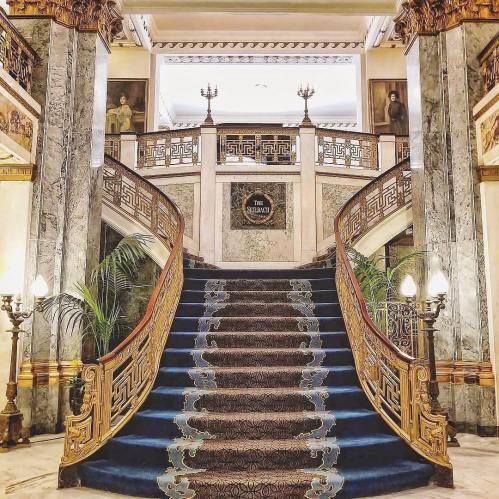 Elegant staircase with ornate railings, blue carpet, and decorative lighting in a grand interior setting.