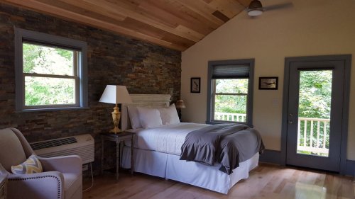 Cozy bedroom with a stone accent wall, a queen bed, armchair, and large windows overlooking greenery.