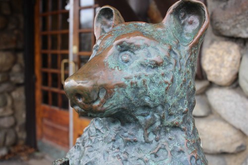 A close-up of a bronze bear statue with a textured surface, set against a stone wall and wooden door.