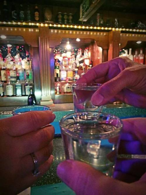 Three hands holding shot glasses in a bar, with a colorful display of bottles in the background.