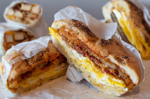 A close-up of a sandwich filled with eggs, bacon, and cheese, with wrapped rolls in the background.