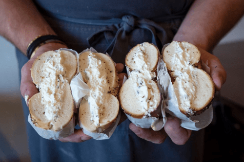 Two bagels filled with cream cheese, held by a person, wrapped in paper.