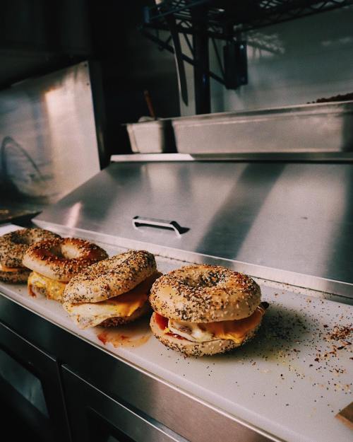 A row of bagel sandwiches with various toppings on a white plate, set against a kitchen background.