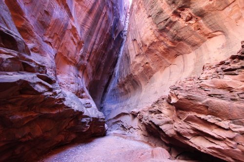 A narrow canyon with towering, colorful rock walls and a sandy floor, illuminated by soft natural light.