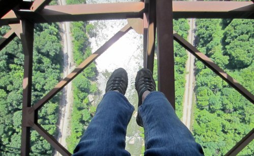 Feet in black shoes dangling over a metal structure, with a view of a river and lush green trees below.