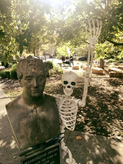 A skeleton poses next to a bust of Washington Irving in a park, surrounded by trees and hay bales.