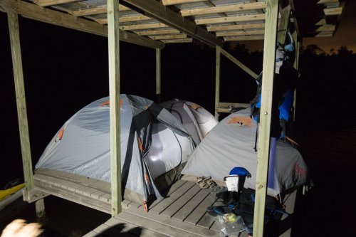Two tents set up on a wooden dock at night, with gear hanging nearby and a dark background of trees.